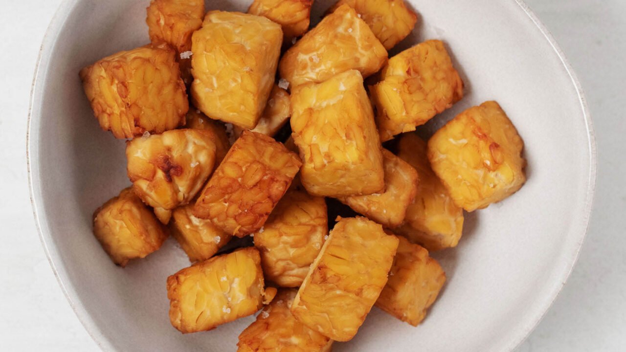 An asymmetrical, ceramic bowl holds browned tempeh "nuggets." It rests on a white surface.